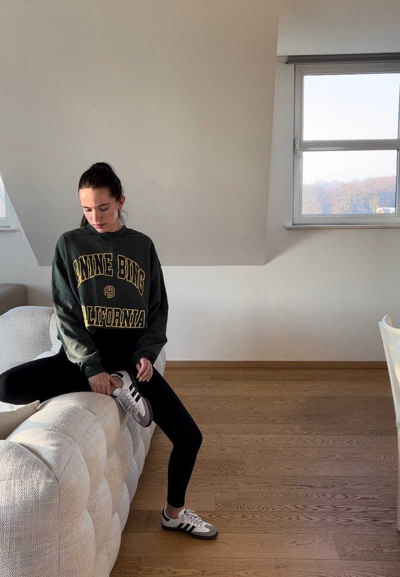 Πράσινο φούτερ με κίτρινα γράμματα, μαύρα κολάν και γκρι αθλητικά παπούτσια με λευκές λεπτομέρειες. Καθισμένο σε έναν υφασμάτινο, ανοιχτόχρωμο καναπέ.