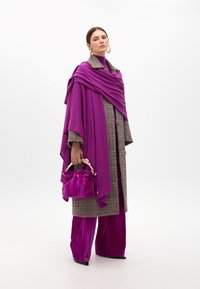 Fioletowy płaszcz, marynarka w kratę oraz żywo fioletowe, plisowane spodnie. Trzyma małą, pluszową welurową torebkę z złotym uchwytem i ściągaczem.