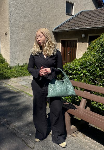 Mono negro de manga larga con pantalones acampanados; bolso tejido verde; entorno al aire libre con un banco y vegetación. Fondo iluminado por el sol.