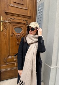 Μαύρο παλτό με υφασμάτινη υφή, μπεζ κασκόλ με φούντες, καφετί καπέλο και σκούρα γυαλιά ηλίου. Ξύλινη πόρτα με περίτεχνα σχέδια στο φόντο.