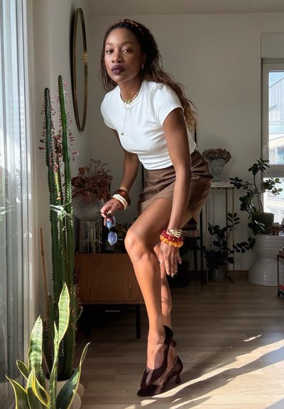 Mujer vestida con blusa blanca, falda marrón y tacones posa en el interior cerca de un cactus alto y una ventana con luz solar y plantas alrededor.