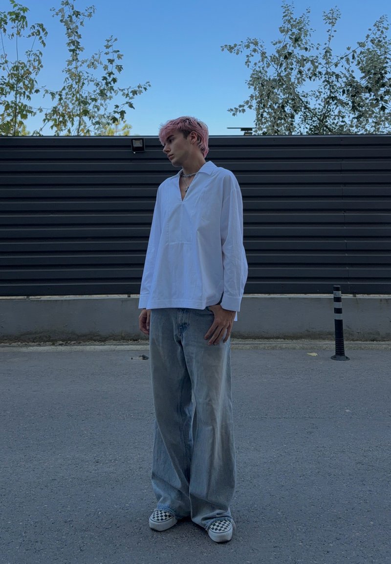 Jeune personne aux cheveux roses se tient sur le trottoir, portant une chemise blanche ample, un jean large et des chaussures slip-on à motif damier près d'une clôture noire avec des arbres.