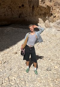 Νεαρό άτομο που χαμογελά, στέκεται σε βραχώδες έδαφος σε έναν φωτεινό από ήλιο σπήλαιο, φορώντας φανελάκι, ριγέ πουκάμισο, σορτς και πράσινα αθλητικά παπούτσια, κρατώντας ένα καπέλο.