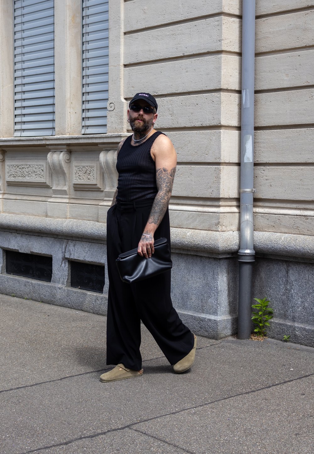 Black ribbed tank top, loose black trousers, green slip-on shoes, and a black clutch bag. Tattooed arm, sunglasses, and a cap worn backwards.