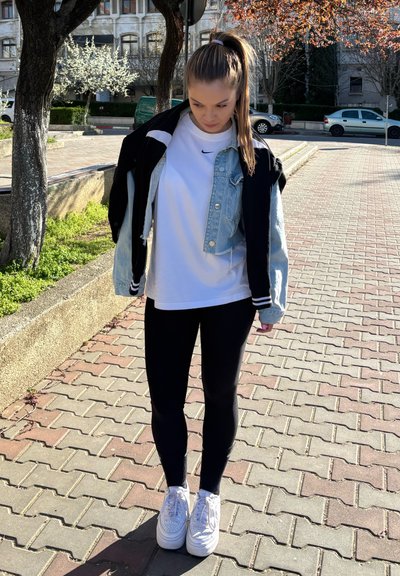 Modelo vistiendo una camiseta blanca, chaqueta de mezclilla y leggings negros; zapatillas blancas con suela texturada, de pie sobre un camino de ladrillos.