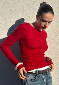 Haut à manches longues rouge en matière légère avec une coupe ajustée. Associé à un jean bleu clair et une ceinture argentée, agrémenté de bracelets épais crème.