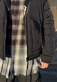 Doudoune noire avec fermeture éclair et cordon de serrage, superposée sur une chemise à carreaux grise et noire avec ourlet effiloché et fermeture à boutons.