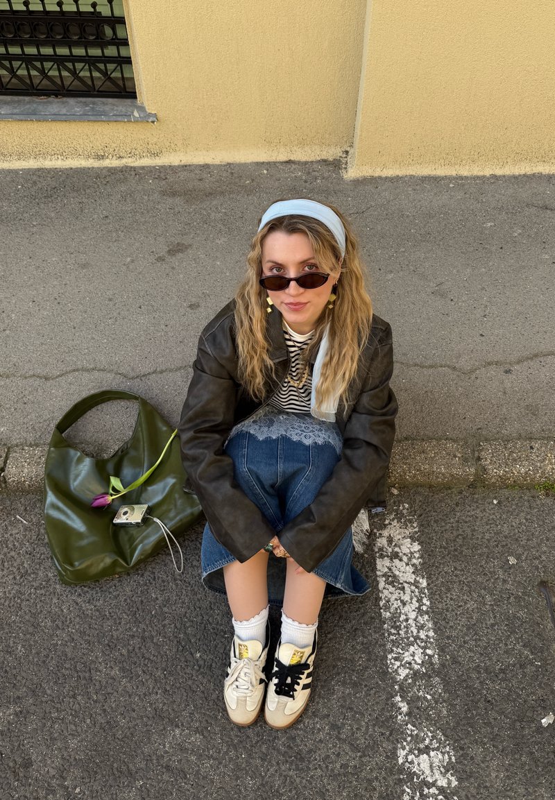 Jeune femme portant des lunettes de soleil, une chemise rayée, une veste en cuir, une jupe en jean, des baskets blanches et un bandeau bleu, assise sur le trottoir à côté d’un sac vert avec une fleur violette.