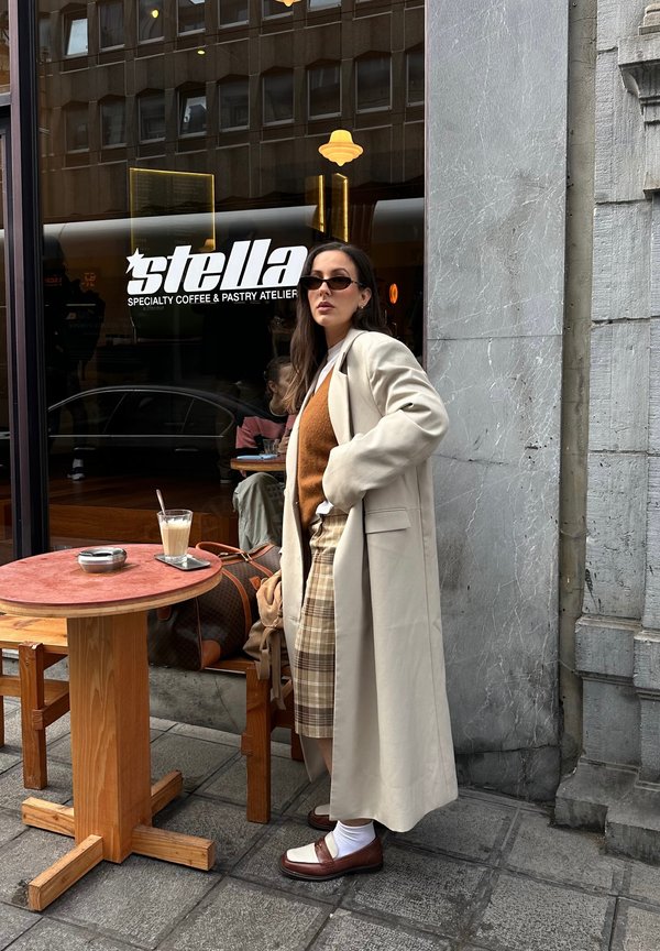 Beige lang frakke over en brun sweater, ternet nederdel og brune sko. Træbord og glas med isdrik synlig i en udendørs caféindstilling.