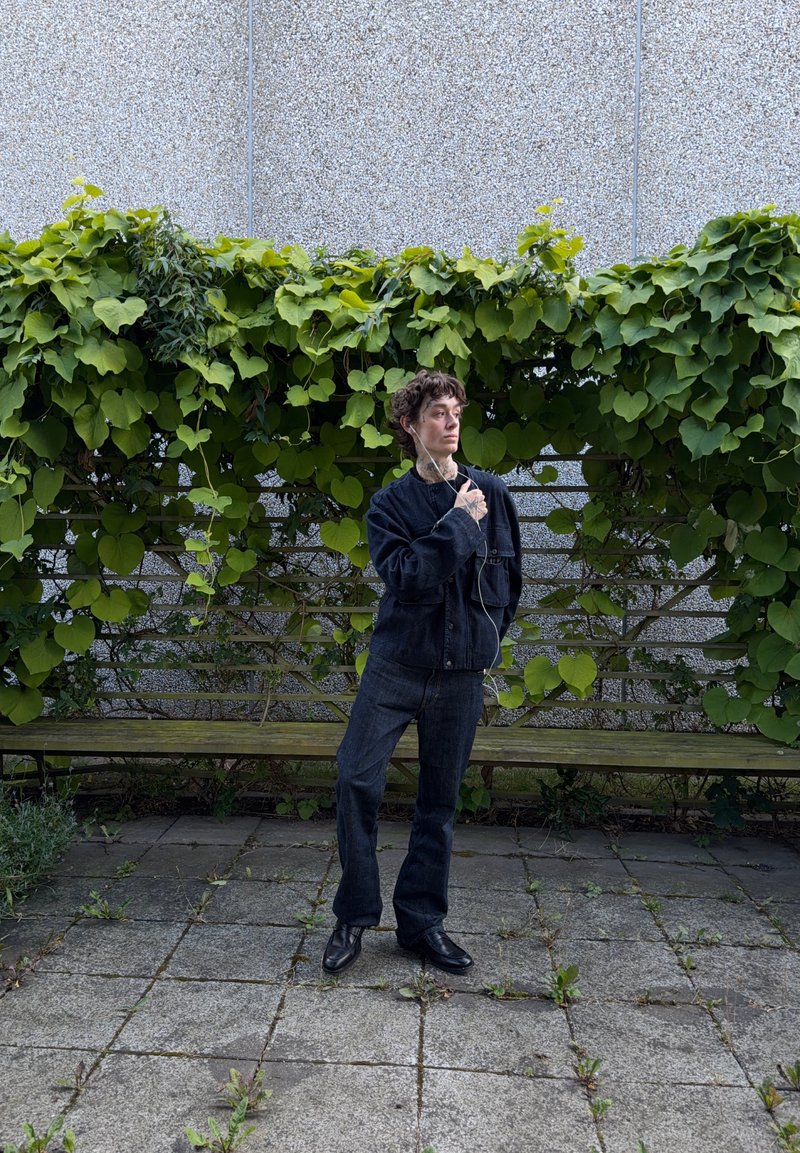Personne aux cheveux courts et bouclés portant une tenue en denim foncé et des chaussures noires se tient sur des dalles en pierre avec un mur recouvert de verdure derrière.