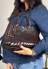 Femme portant un cardigan bleu tenant un grand sac à main en cuir tressé brun foncé avec des poignées courtes sur un fond clair.