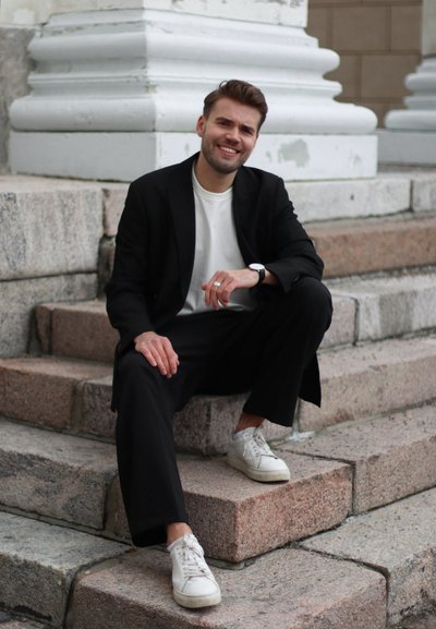 Blazer negro y pantalones llevados sobre una camiseta blanca. Sentado en escalones de piedra, con zapatillas blancas, y el cabello peinado en suaves ondas.