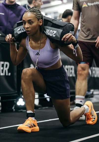 Atleta con trenzas realiza una zancada con una bolsa de peso negra. Lleva un top corto lila claro, pantalones cortos oscuros y zapatos naranjas.