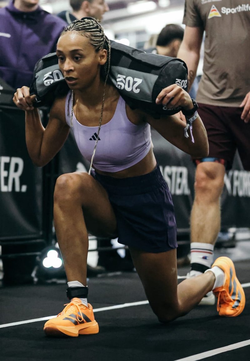 Une athlète aux cheveux tressés effectue une fente avec un sac lesté noir. Elle porte un crop top violet clair, un short foncé et des chaussures orange.