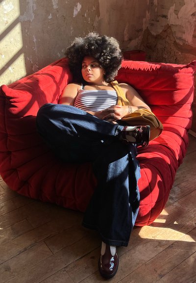 Sillón grande y de felpa de color rojo con un diseño acolchado. Persona reclinada, vistiendo una camiseta de tirantes a rayas y jeans de campana, con zapatos marrones.