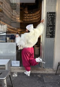 Femme en veste blanche pelucheuse et pantalon rouge, debout sur une jambe tenant une tasse de café à l'extérieur d'un café avec un tableau de menu accroché au mur.