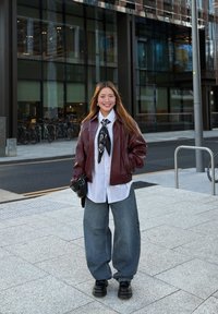 Bordó bőrkabát, fehér ing, fekete selyemsál, laza szürke farmernadrág, fekete platform cipők; mosolyogva, hosszú hajjal a szabadban.