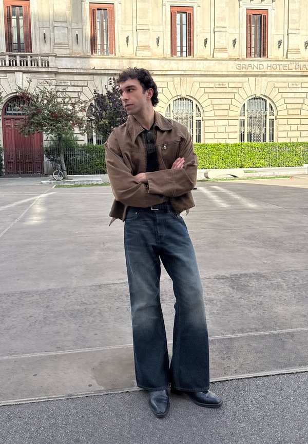 Smeđi skraćeni kaput preko crne majice, uparen s širokim blijedoplavljenim trapericama i crnim cipelama. Glatka tekstura i ležeran dizajn.