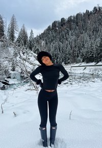 Czarna długa bluzka i legginsy w połączeniu z szarymi wodoodpornymi butami, na tle zaśnieżonego krajobrazu i drzew iglastych w tle.
