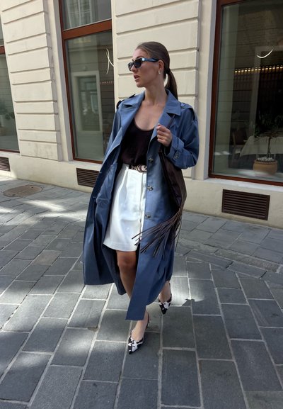Mujer caminando por una acera pavimentada con un abrigo azul tipo gabardina, falda blanca, top negro, zapatos de tacón con estampado de vaca, gafas de sol y llevando un bolso negro con flecos.