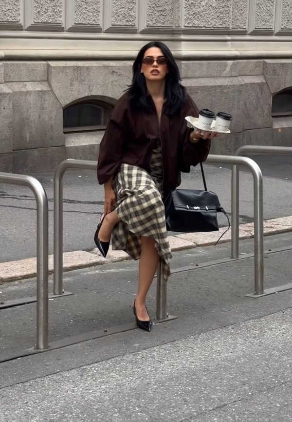 Mujer con gafas de sol y falda de cuadros sentada en una barandilla de metal sosteniendo una bandeja con dos tazas de café en una calle de la ciudad.