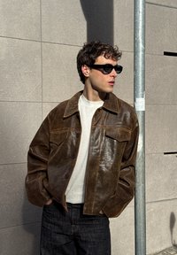 Bruine leren jas met een licht glanzende afwerking, voorzien van een puntige kraag en voorzakken, gecombineerd met een witte trui en donkergroene jeans.