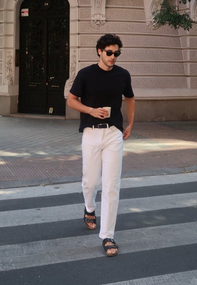 Camiseta negra, pantalones blancos y sandalias oscuras. Sosteniendo una taza de café mientras camina por un paso de peatones en una calle de la ciudad. Edificio al fondo.