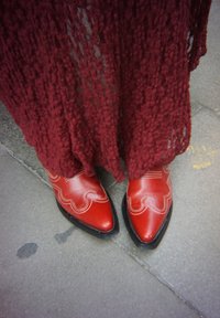 Červené kožené čižmy so špicatou špičkou, ozdobené vlnkovaným vzorom a bielym prešitím, v kombinácii s dlhým bordovým pleteným odevom s textúrou.