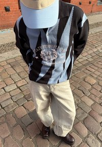 Raidallinen musta ja vaaleansininen neulottu pusero logolla, beige lippalakki, vaaleat leveälahkeiset housut ja tummat kengät mukulakivikadulla.