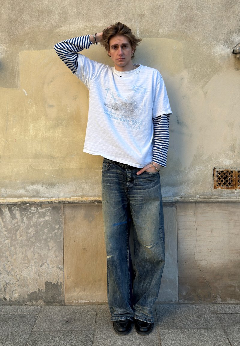 Jeune homme avec la main sur la tête portant un t-shirt à manches longues rayé superposé et un t-shirt blanc graphique, jean ample, debout contre un mur beige texturé.