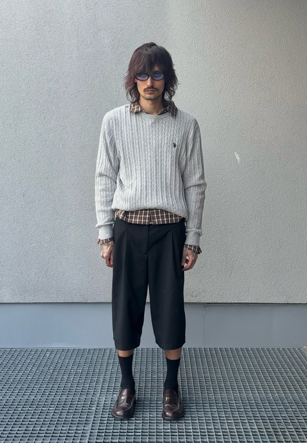 Homme aux cheveux mi-longs porte des lunettes de soleil bleues, un pull gris à torsades par-dessus une chemise à carreaux, un pantalon noir court, des chaussettes foncées et des mocassins marron.