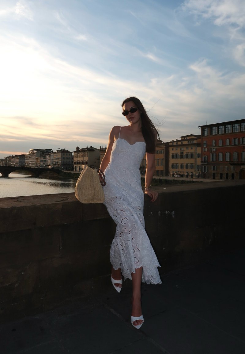 Femme en robe blanche et lunettes de soleil appuyée sur un mur en pierre au bord de la rivière au coucher du soleil, avec des bâtiments historiques et un pont en arrière-plan.