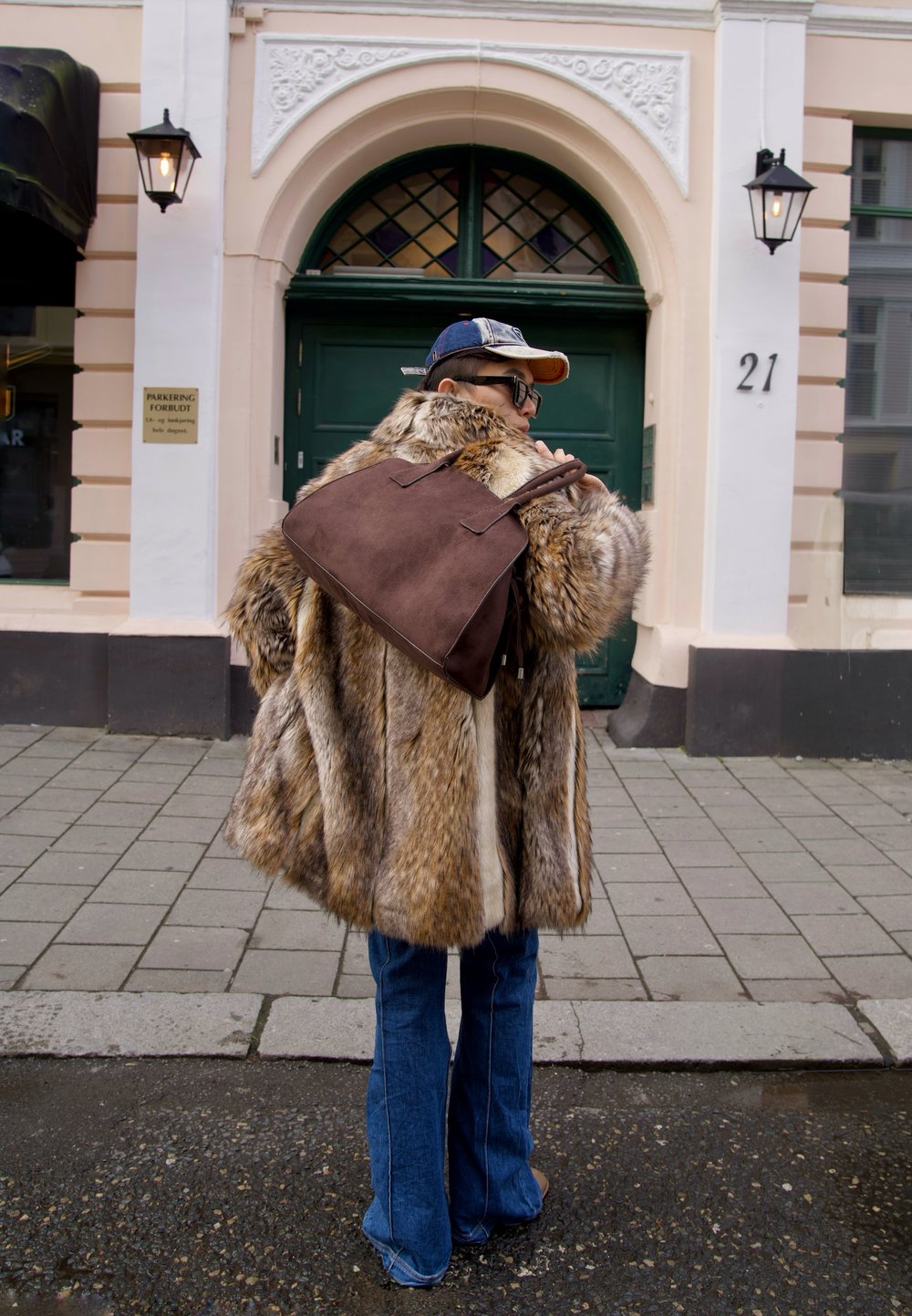 Kabát z umělé kožešiny v béžových a hnědých tónech, doplněný rozšířenými džínami. Hnědá semišová taška přes rameno na historickém pozadí.