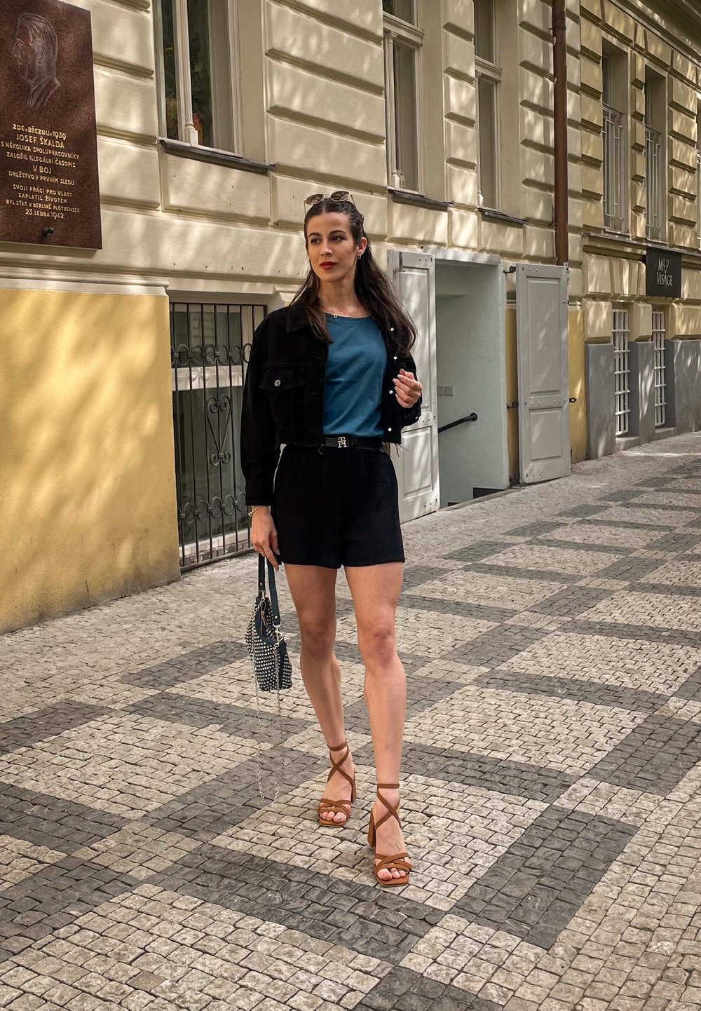 Melnas denim jaka, tirkīza tops, melni šorti un brūni siksnu sandaļi. Tur rokās studētu somu, stāvot uz rakstainām akmens bruģa.