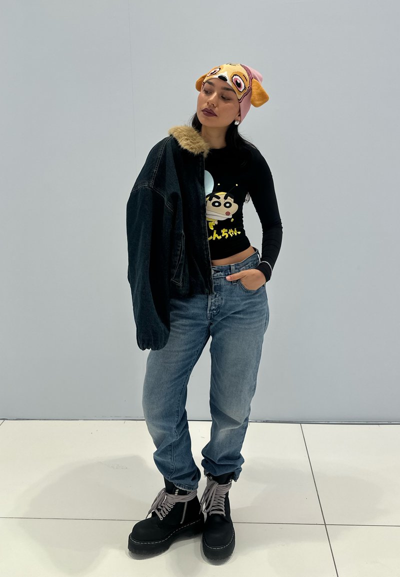 Femme portant un bonnet cartoon et un crop top noir à manches longues avec un personnage animé, un jean ample, des bottes noires, et une veste à col en fourrure posée sur une épaule.