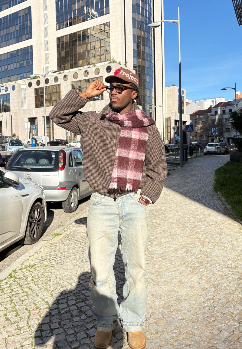 Pull surdimensionné marron à carreaux, écharpe à carreaux rouge et blanc, jeans bleu clair déchirés, et bottes beige; lunettes de soleil et casquette complètent le look.