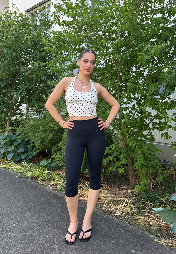 Ung kvinde med mørkt hår iført en hvid crop top med prikker, sorte leggings og flipflops, der står ude nær grønne buske.