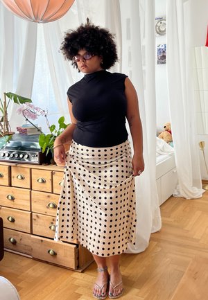 Top sin mangas negro, falda beige con lunares, sandalias planas; cómoda de madera y planta en el fondo; luz natural de la ventana.
