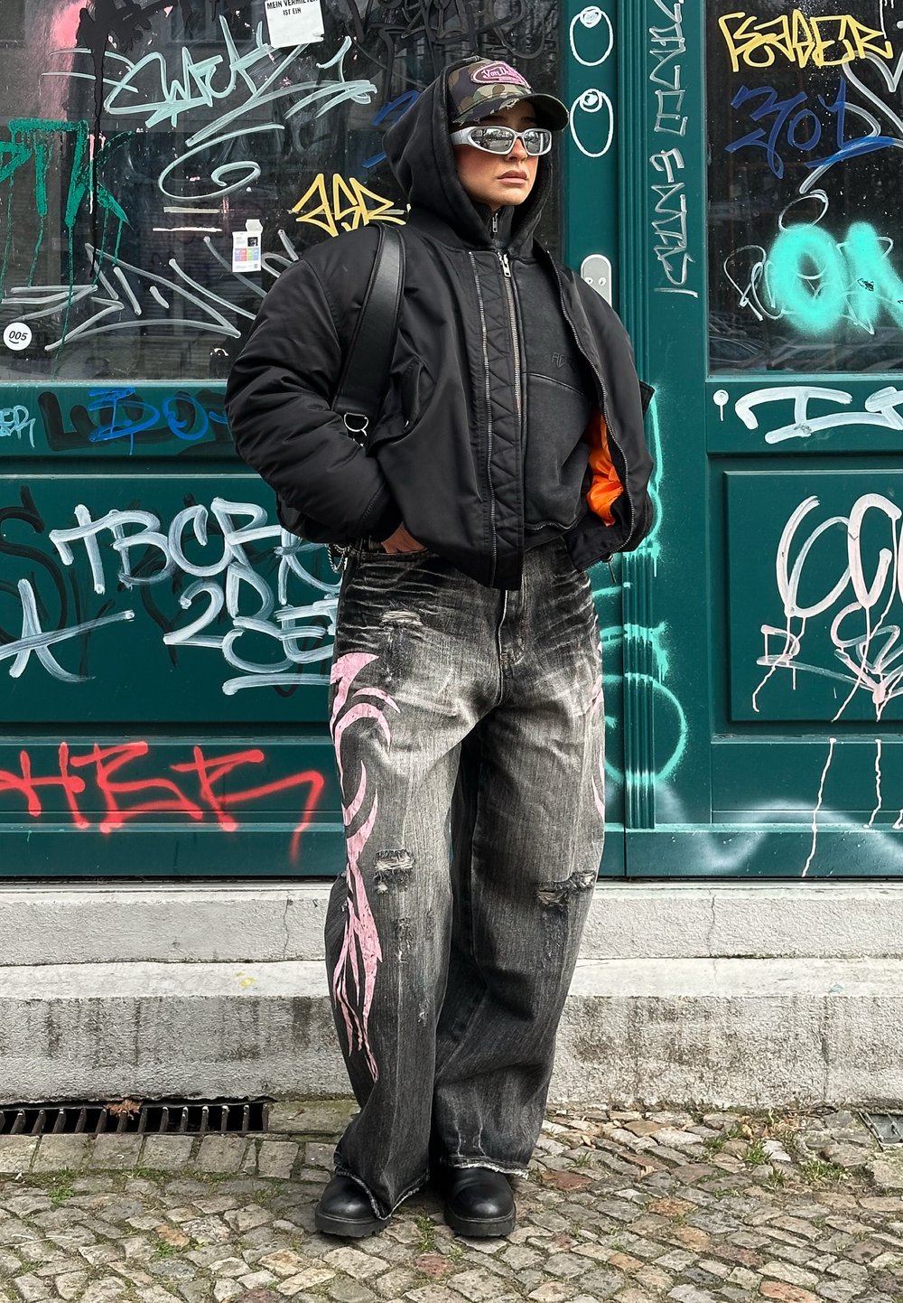 Μαύρο παλτό με επένδυση πάνω από γκρι φούτερ, κατεστραμμένο μαύρο τζιν με ροζ σχέδια και μαύρες μπότες. Αξεσουάρ περιλαμβάνουν γυαλιά ηλίου και καπέλο.