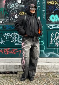 Μαύρο παλτό με επένδυση πάνω από γκρι φούτερ, κατεστραμμένο μαύρο τζιν με ροζ σχέδια και μαύρες μπότες. Αξεσουάρ περιλαμβάνουν γυαλιά ηλίου και καπέλο.