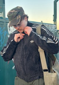Black, fuzzy zip-up jacket with white stripes on the sleeves and Adidas logo; paired with a camo cap and a beige shoulder bag.
