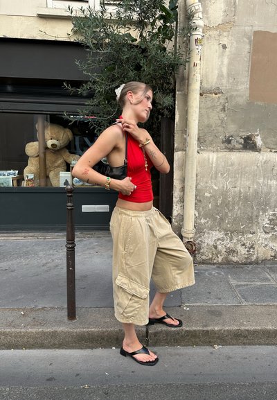 Top corto rojo de halter, pantalones cargo anchos beis con bolsillos y chanclas negras. De pie junto a una pared con un oso de peluche detrás de un cristal.