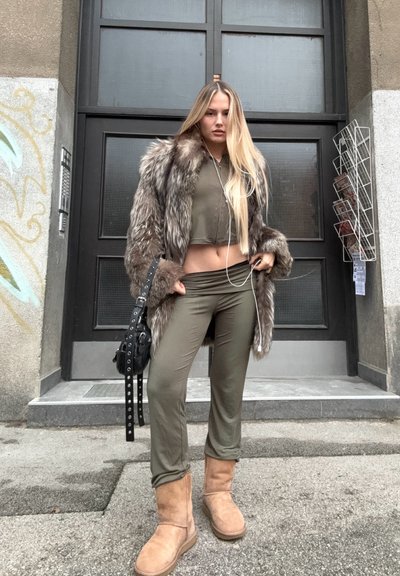 Abrigo de pelaje sintético marrón, top corto verde oliva, pantalones a juego y botas de rodilla color crema. Bolsa de hombro negra con acentos metálicos.