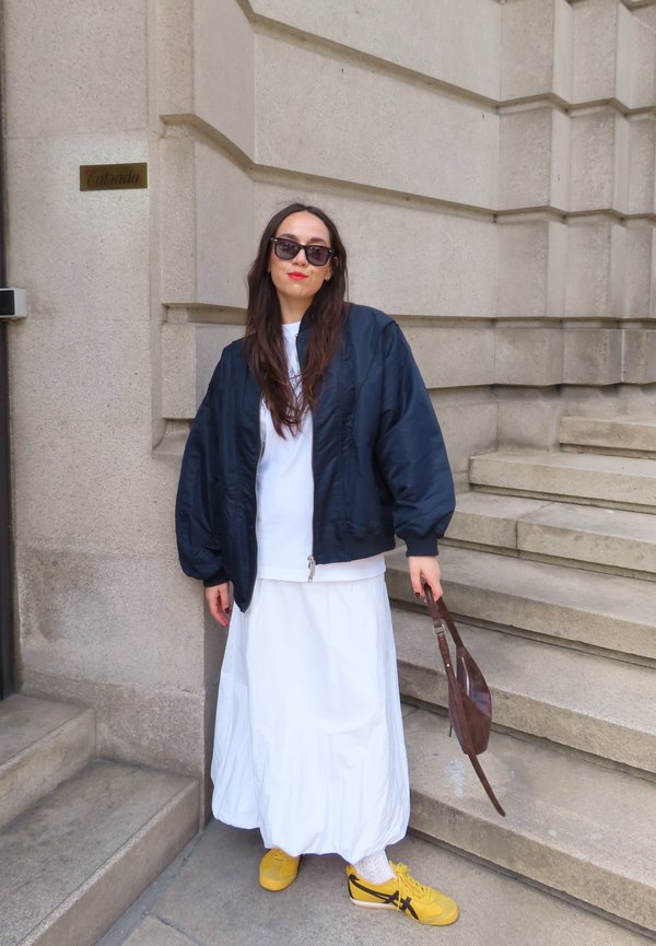 Femme en veste noire, robe blanche, baskets jaunes et lunettes de soleil, debout près d'un escalier en pierre, tenant un sac marron près de l'entrée d'un bâtiment.