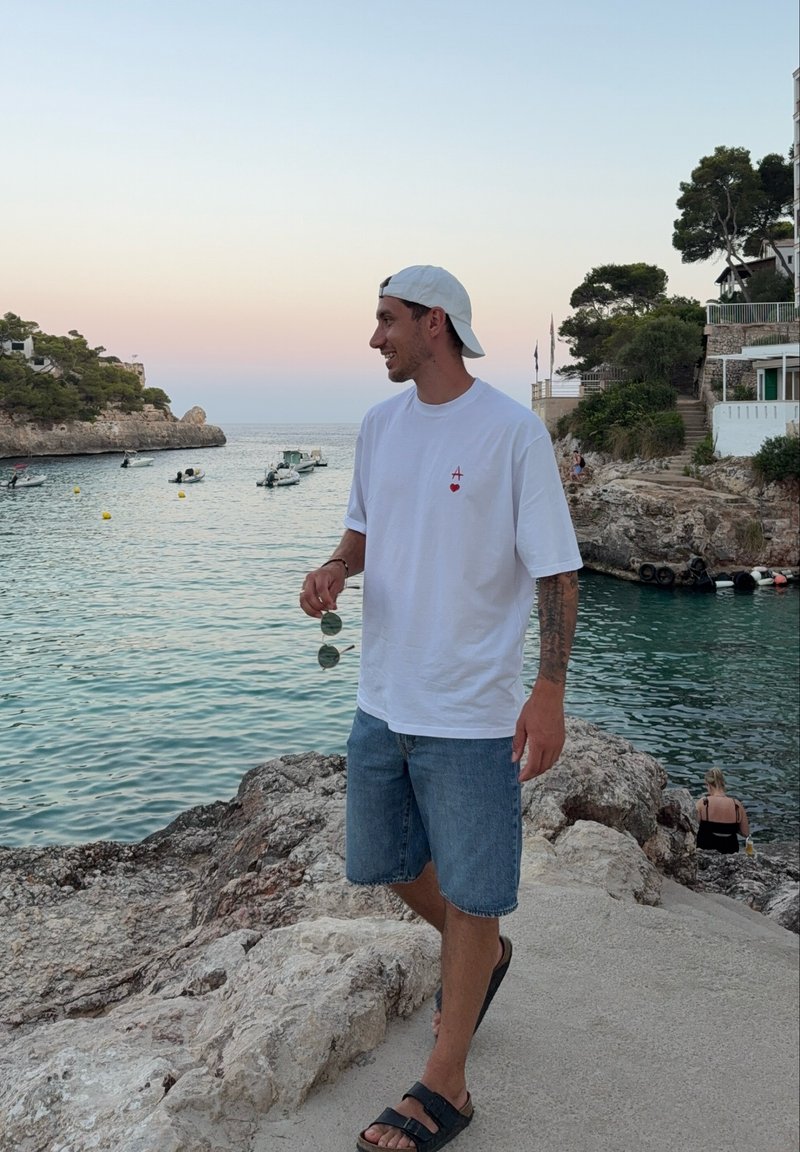 Νέος άνδρας με λευκό καπέλο, λευκό μπλουζάκι και τζιν σορτς στέκεται σε βραχώδη ακτή δίπλα σε ήρεμη θάλασσα με βαρκούλες και δέντρα στο ηλιοβασίλεμα.