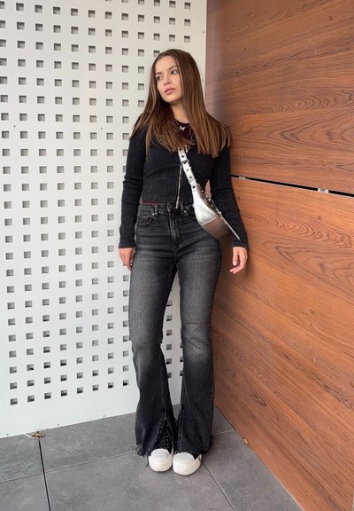 Chaqueta negra corta sobre una blusa burdeos, combinada con jeans negros de campana con aberturas laterales, y zapatillas blancas. Un bolso plateado cruzado complementa el atuendo.