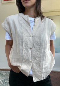Vesta din in de culoare bej deschis, cu mâneci scurte, având închidere cu butoane în față și accente simple cusute, purtată peste un tricou alb.