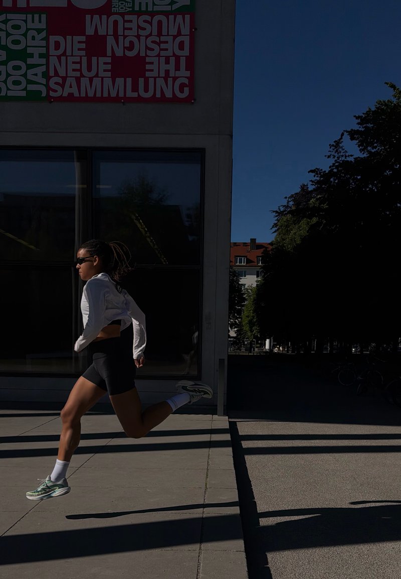 Personne athlétique avec des lunettes de soleil courant rapidement sur un trottoir ensoleillé à côté d'un bâtiment avec une signalisation colorée et des arbres en arrière-plan.