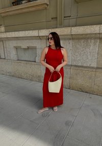 Robe sans manches rouge, longueur cheville avec un détail noué ; tenant un sac à main crème ; lunettes de soleil ; chaussures plates de couleur claire ; fond de mur en pierre.