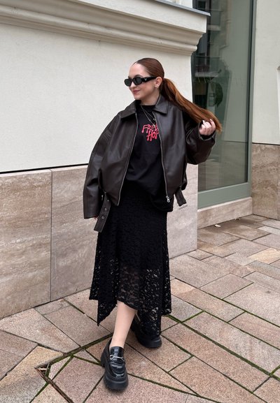 Mujer con una coleta larga que lleva gafas de sol, chaqueta de cuero negra, camiseta gráfica negra, falda de encaje y zapatos de plataforma negros, de pie sobre el pavimento.